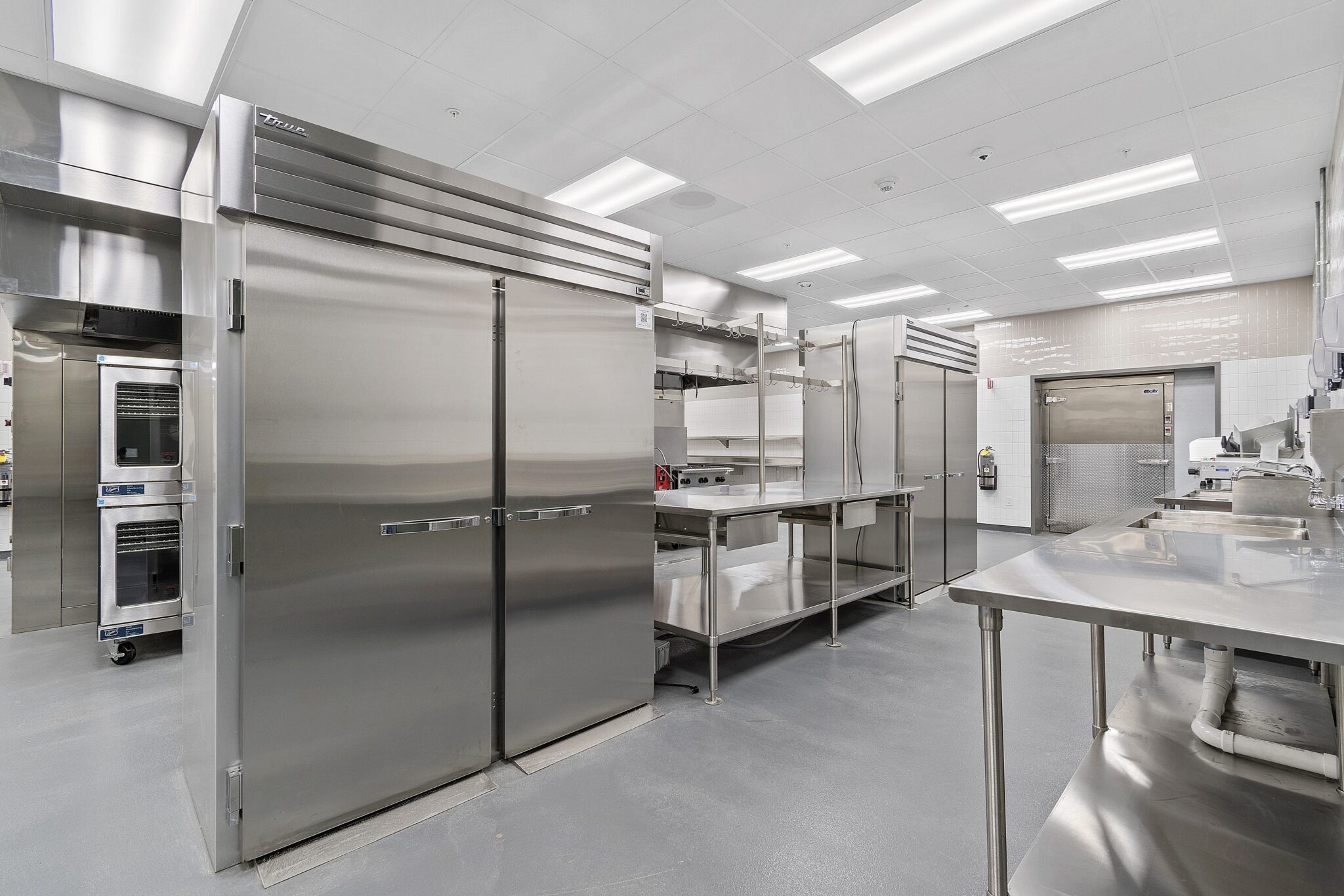 A modern kitchen featuring stainless steel refrigerators, open shelving, countertops, and a clean, organized layout. Bright, spacious environment.