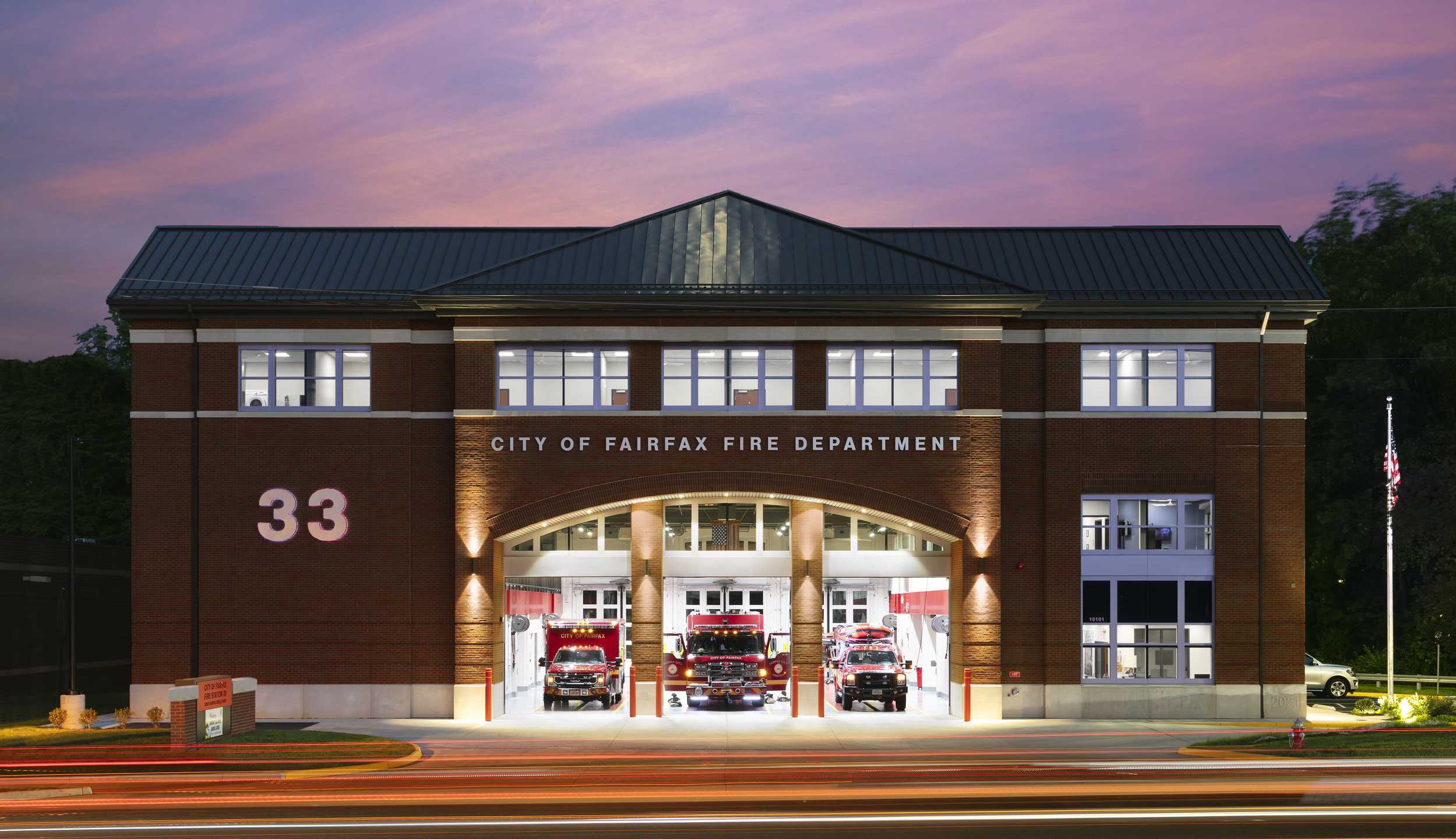 A modern fire department building with illuminated bay doors, featuring fire trucks and a welcoming entrance, set against a twilight sky.