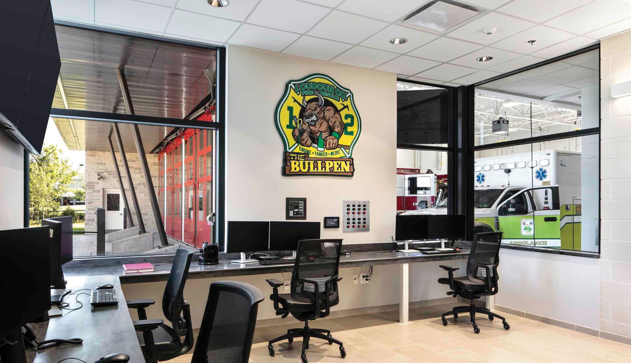 A modern fire station office and watch room features sleek desks and chairs, large windows with a view of emergency vehicles, and a vibrant wall mural.