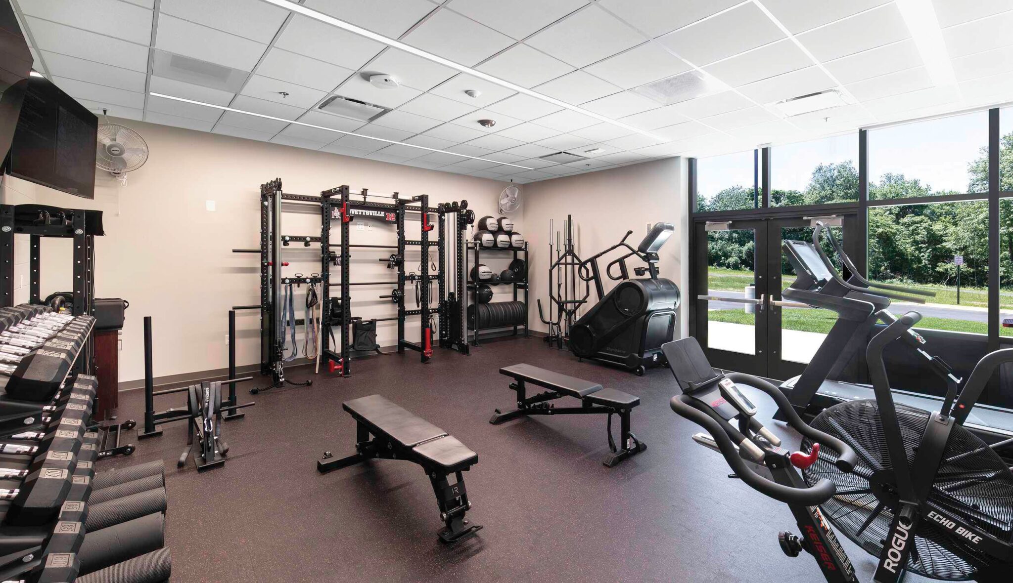 A modern fitness room with weight racks, benches, cardio machines, and large windows providing natural light and views of greenery outside.