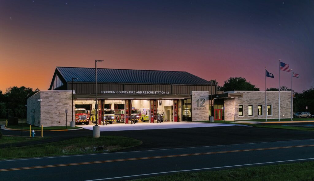 A modern fire station at dusk, featuring bi-fold bay doors, fire trucks, and flags, set against a tranquil evening sky.