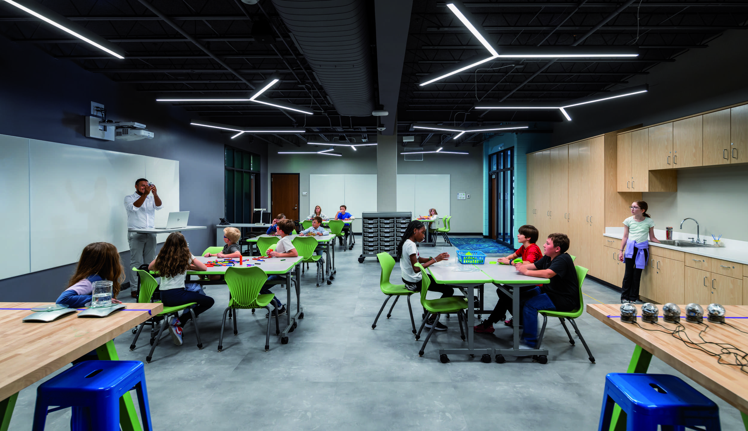 A modern classroom features flexible seating with students engaged in various activities, bright natural light, and interactive learning spaces.
