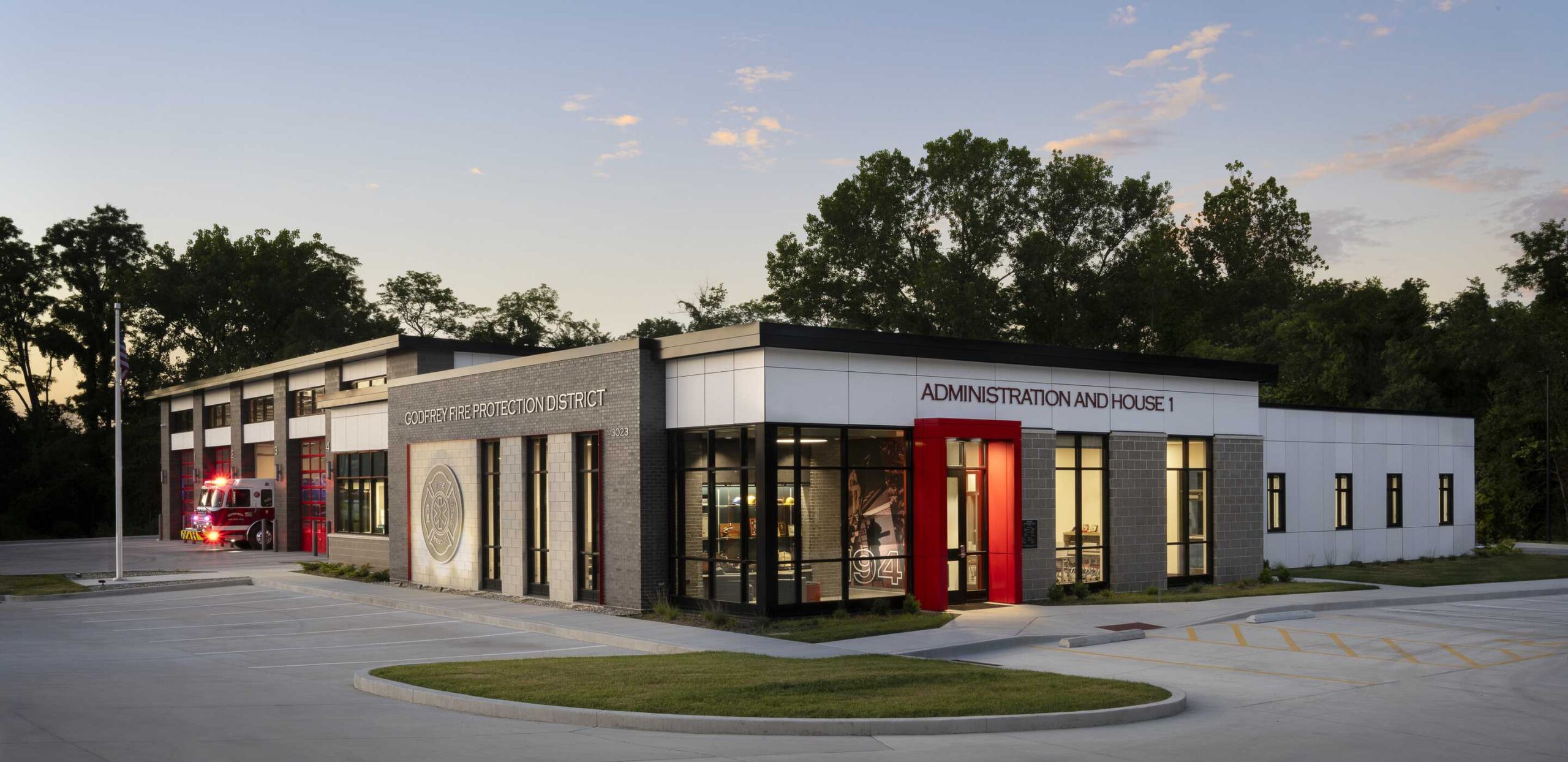 A modern two-story building with a striking design features large windows, red accents, and a landscaped parking area, set against a wooded backdrop.