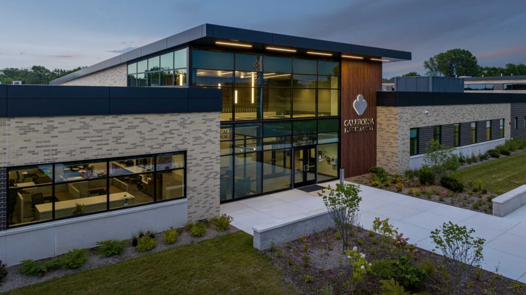 A modern building features large glass windows and a prominent wooden sign, surrounded by landscaped greenery. It's inviting and contemporary.