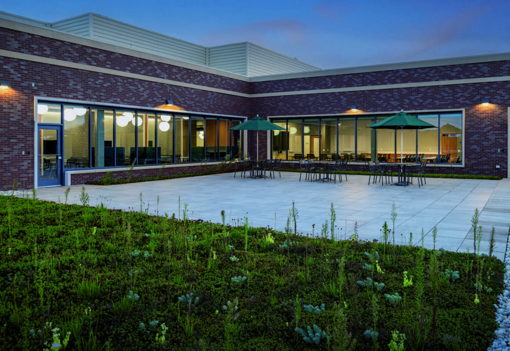 A modern courtyard features stone flooring, outdoor seating under green umbrellas, and lush, low-maintenance landscaping, set against a brick building.