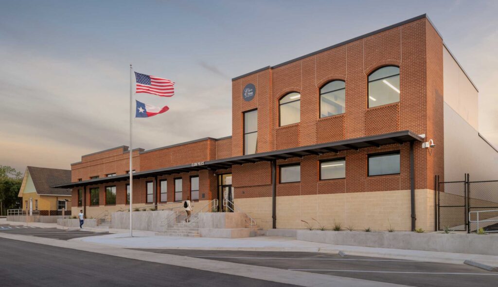 A modern brick building with large windows, featuring an American and Texas flag, set against a serene sky. People walk nearby.