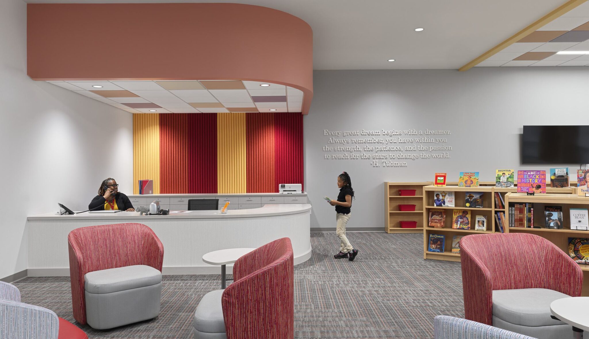 A bright, welcoming reception area features a curved desk, colorful wall panels, cozy seating, and a nearby bookshelf filled with children's books.