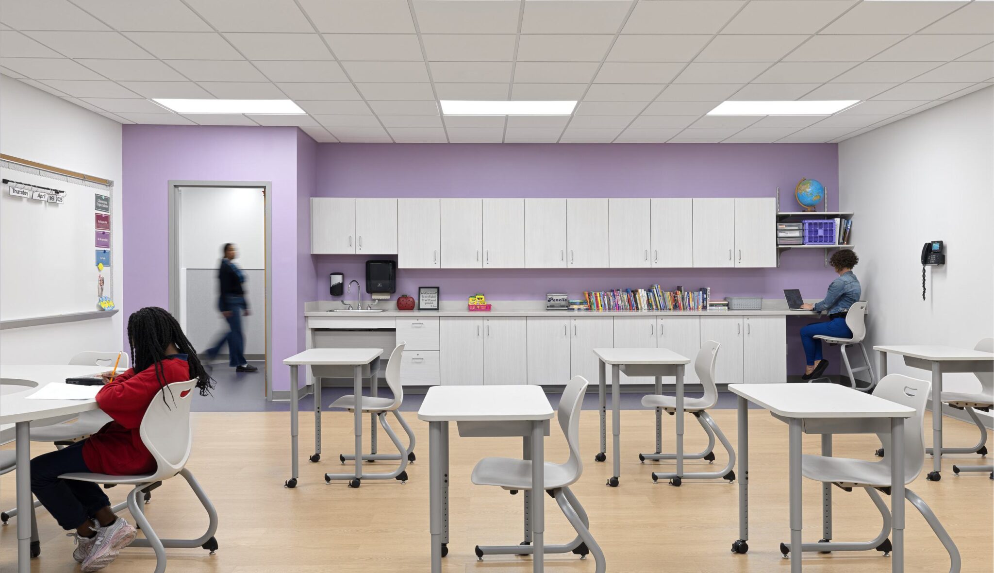 A modern classroom featuring desks, a book storage area, and two students engaged in different activities, with a calming purple backdrop.