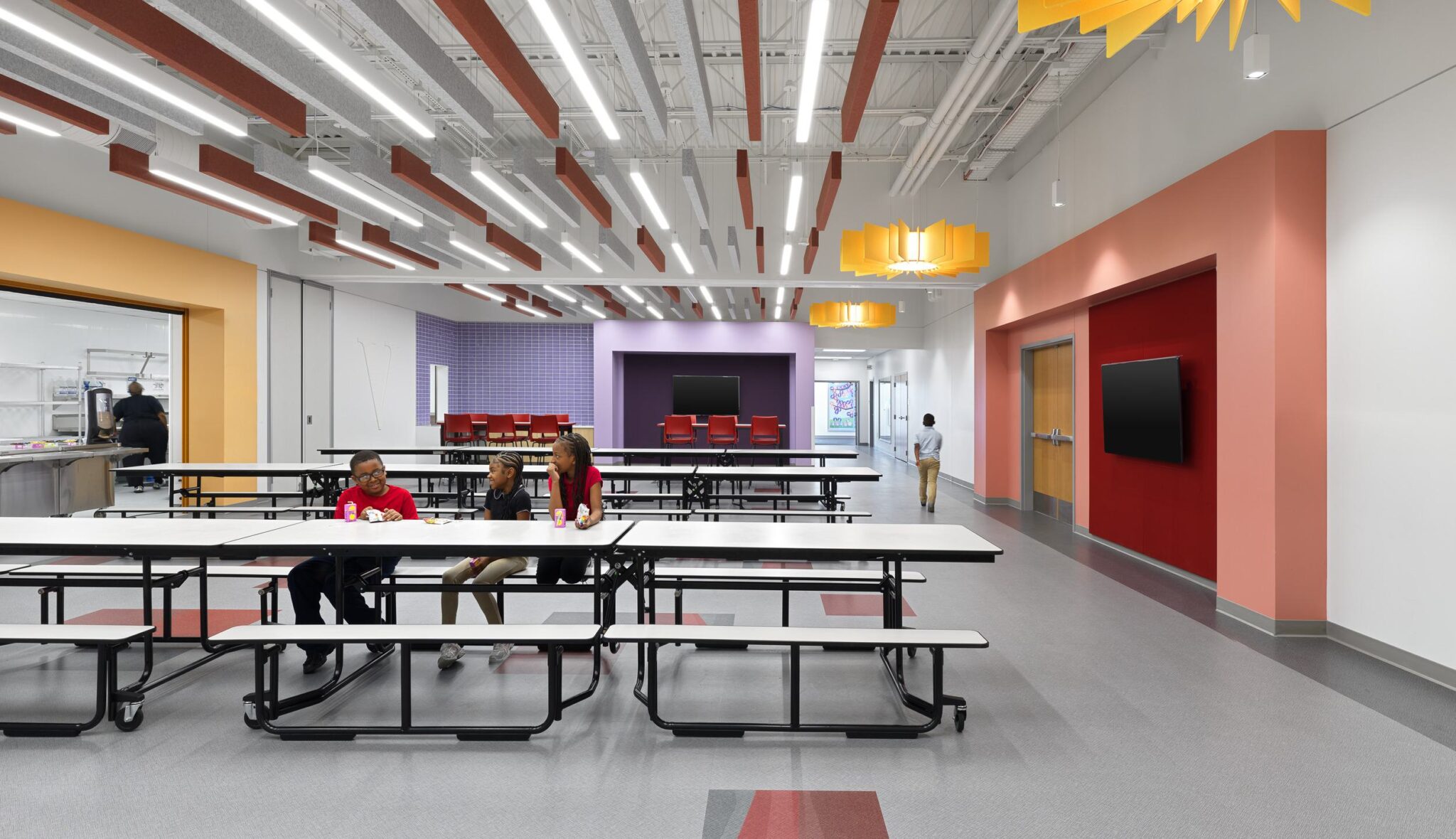 A bright cafeteria features modern design elements, colorful accents, and several long tables, with students seated and engaging.