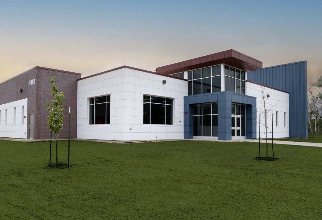 A modern building with a mix of white and blue panels, large windows, and a red roof, situated on a grassy landscape.