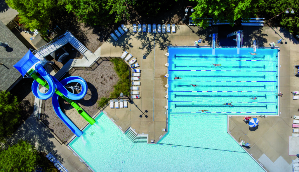 Aerial view of a vibrant water park featuring a racing slide and several swimming lanes, surrounded by lounge chairs and lush greenery.