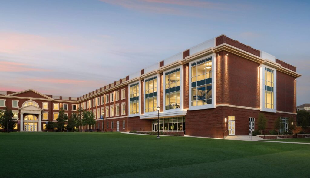 A modern brick building with large windows stands amidst green landscaping, illuminated under a soft twilight sky, showcasing a serene atmosphere.
