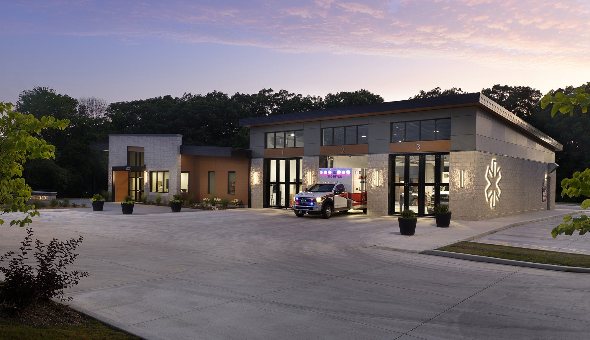 A modern emergency services building features large windows, an ambulance at the entrance, and a landscaped exterior in a serene setting.
