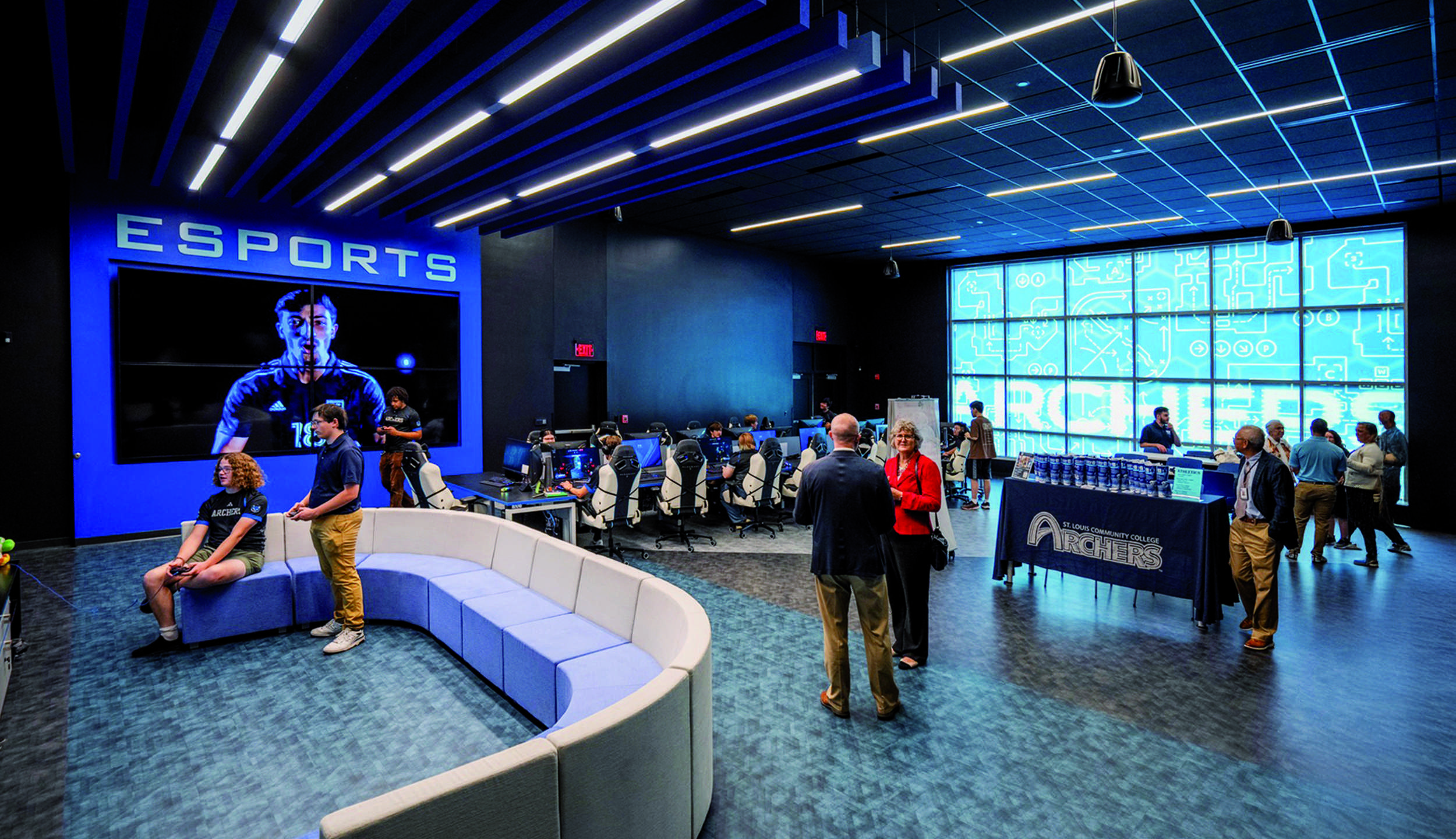 A modern esports arena featuring a large screen, gaming stations, and a sleek, blue-lit design. People are engaged in gaming and conversation.