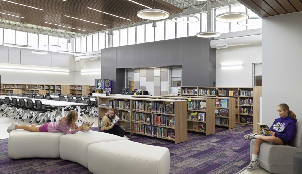 A modern library features students reading on furniture, surrounded by shelves of books, with a spacious, well-lit design.