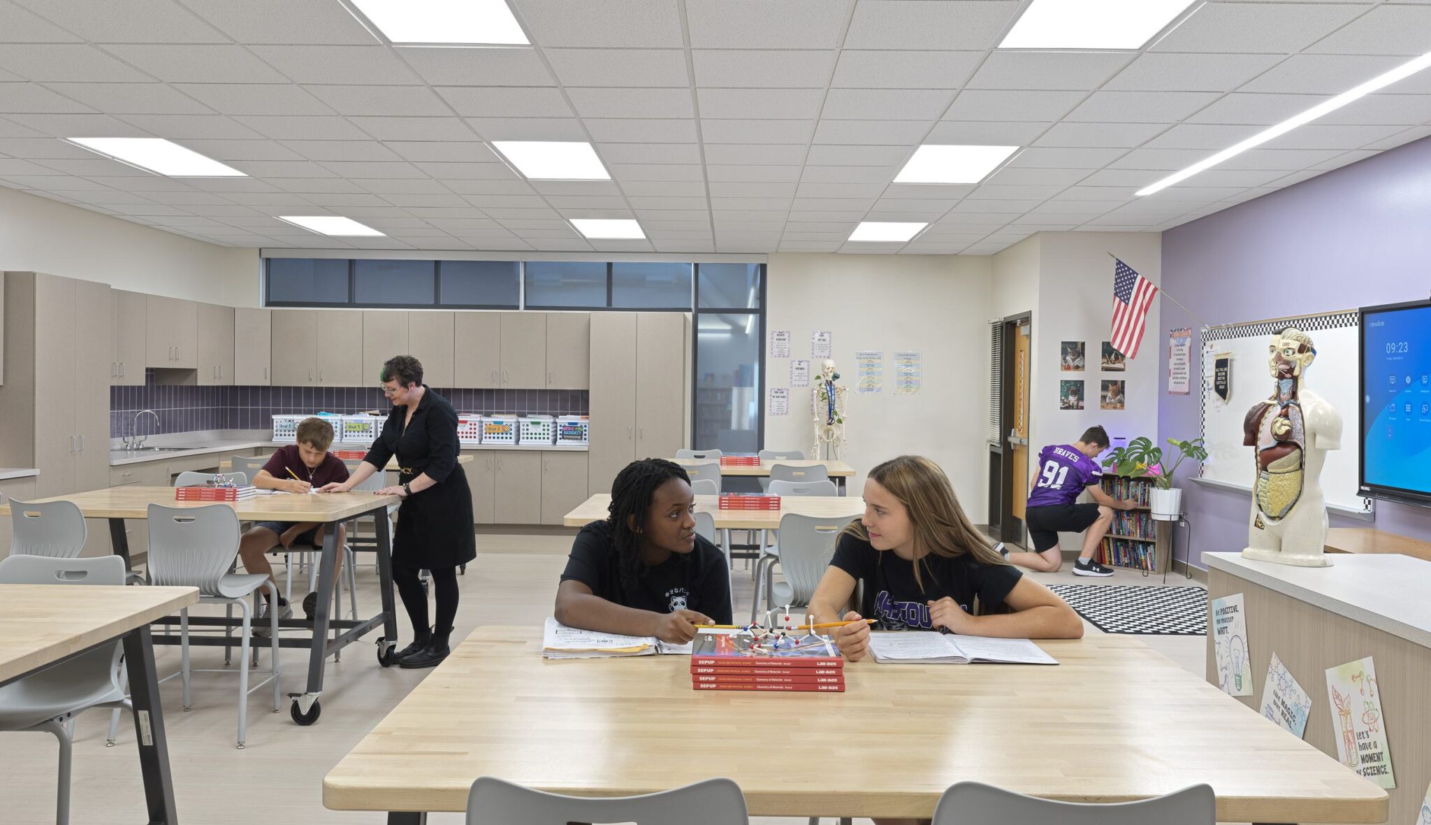 Students collaborate at tables in a modern classroom, while a teacher assists and others engage with resources in a bright, lively environment.
