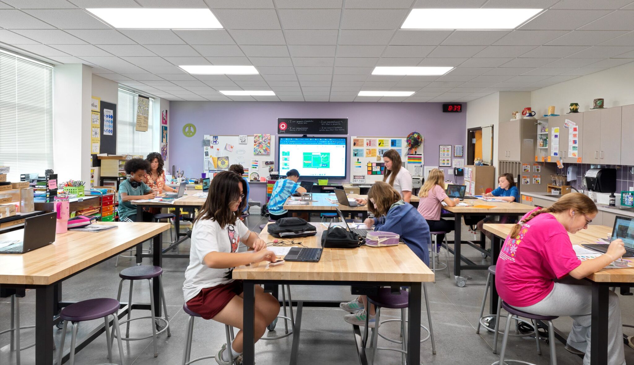 A classroom filled with students working on laptops, engaged in learning, with a teacher providing assistance and a digital board displaying content.