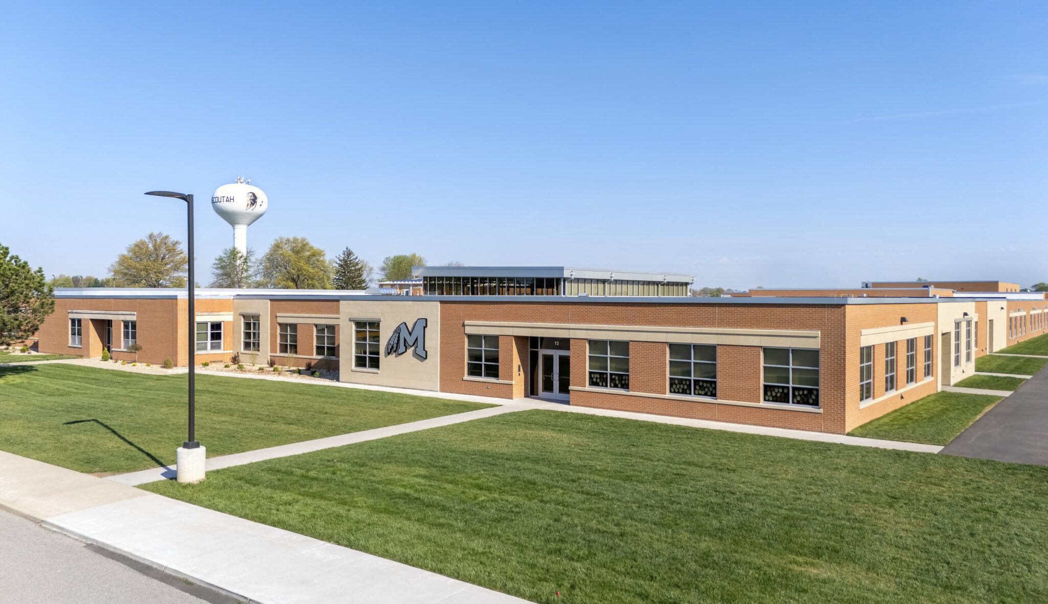 A modern school building with a prominent "M" logo, surrounded by green grass and trees, features a water tower nearby against a clear sky.