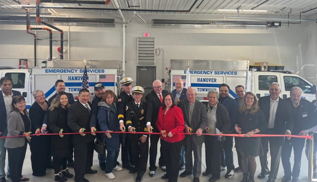 A diverse group gathers to cut a ceremonial ribbon in front of emergency service vehicles, celebrating a new facility.