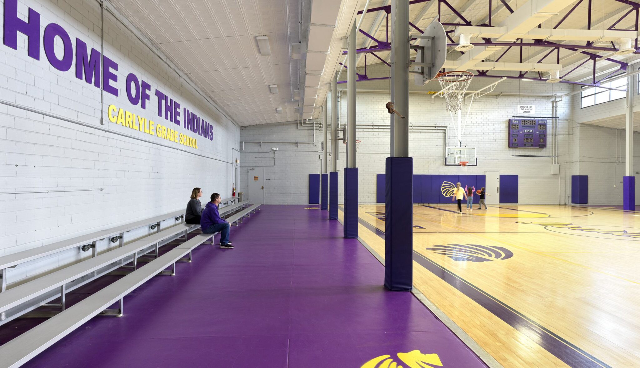 A gymnasium features wooden basketball floors, purple padding along the walls, and bleachers with a few people seated.