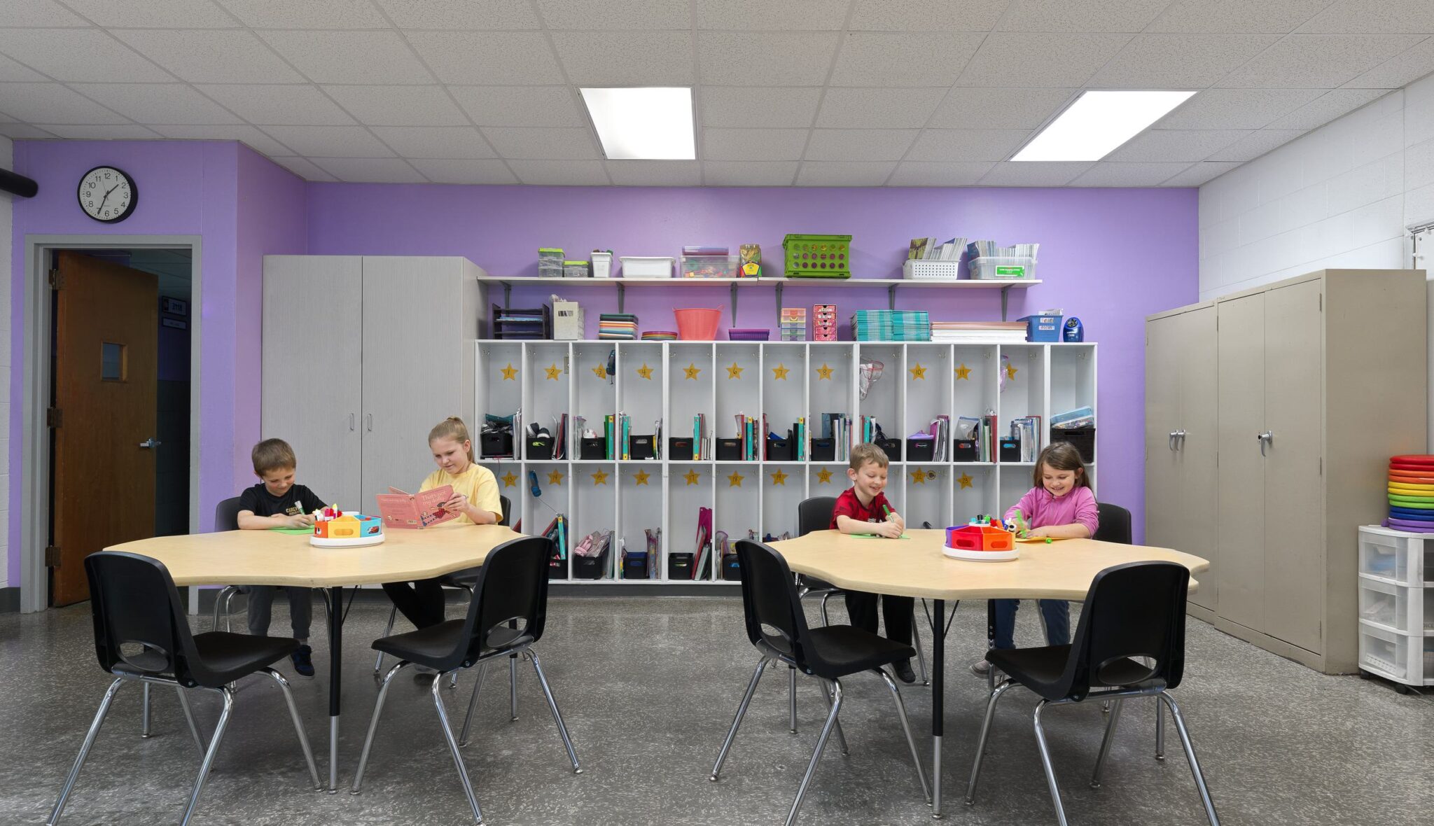 A colorful classroom features four children engaged in activities at a large table, with shelves of supplies and a clock on the wall.