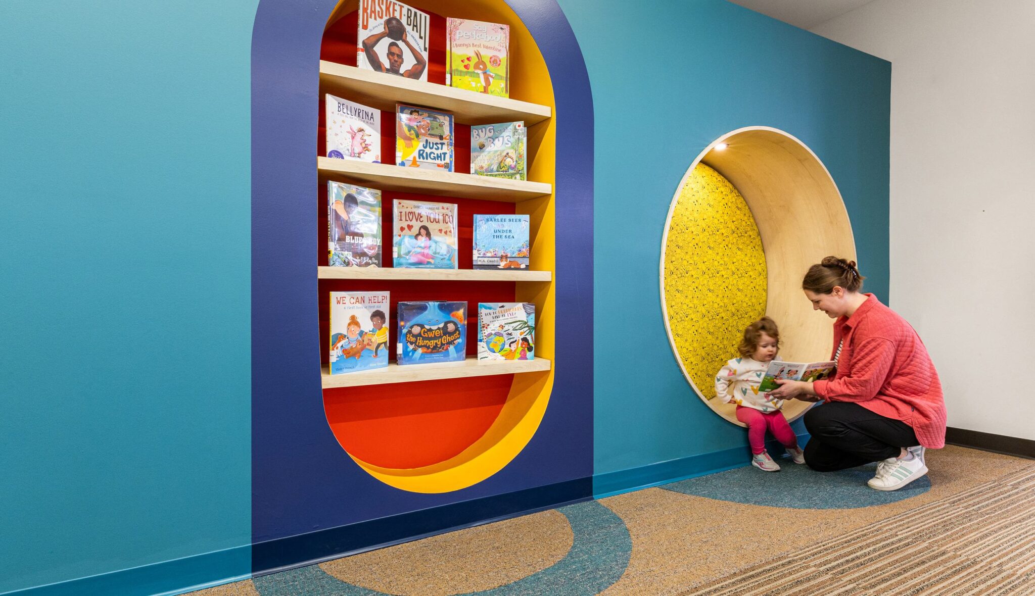 A colorful reading nook features a book shelf and a cozy space where a child and caregiver engage with a story together.