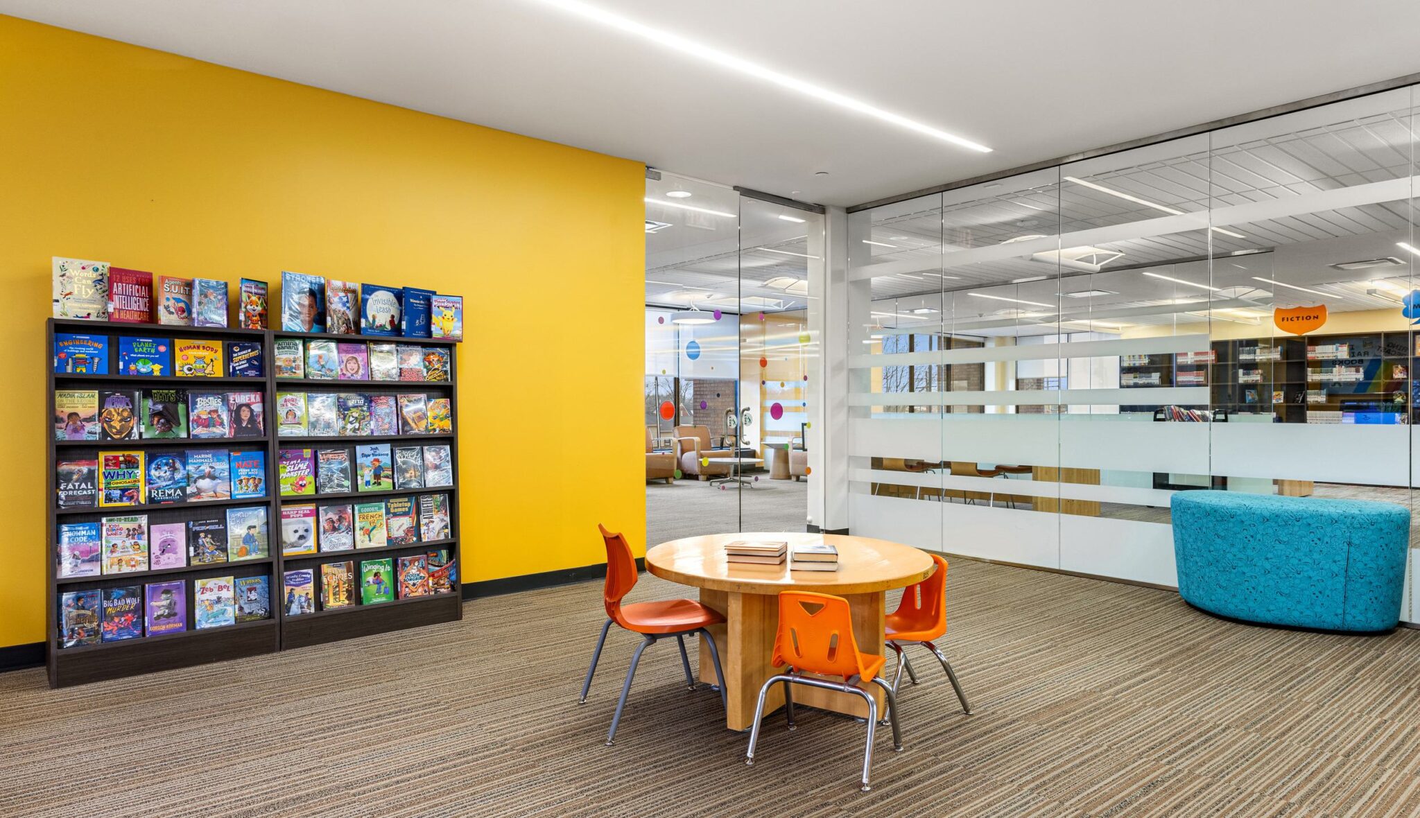 A bright, welcoming space features a colorful book display, round table with chairs, and cozy seating against a vibrant yellow wall.