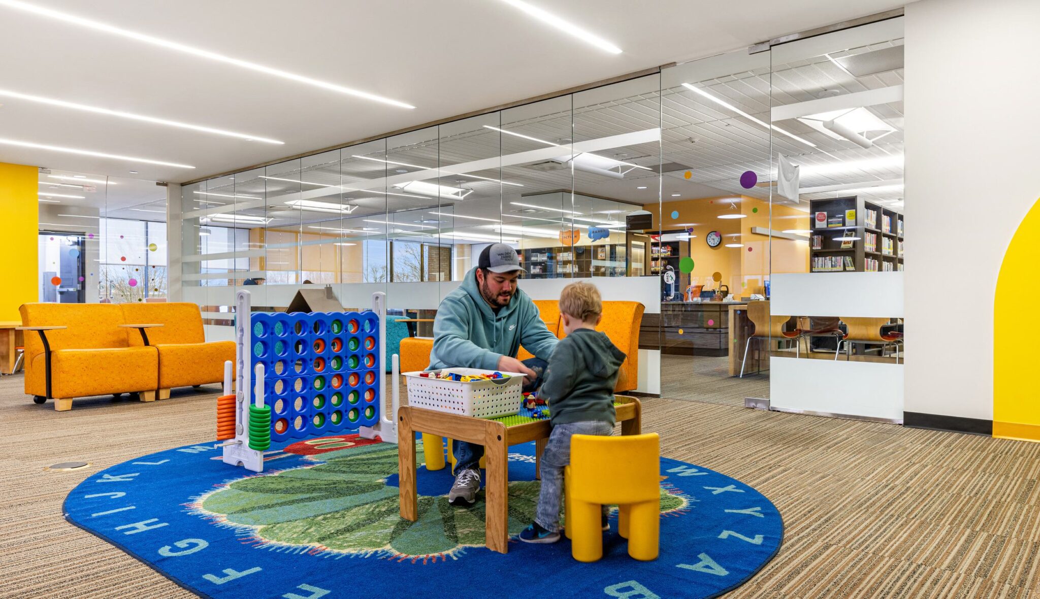 A man and a child play together at a colorful, modern library with comfy seating and bright decor. Educational toys are scattered nearby.