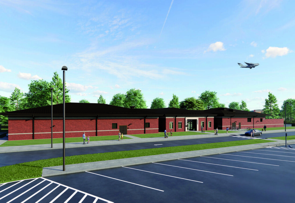A modern red-brick building with large windows, surrounded by green trees, parked cars, and an airplane flying overhead.