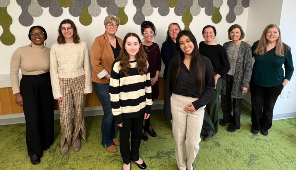 A diverse group of women stands together, smiling in a bright room with green carpet and an artistic wall design.