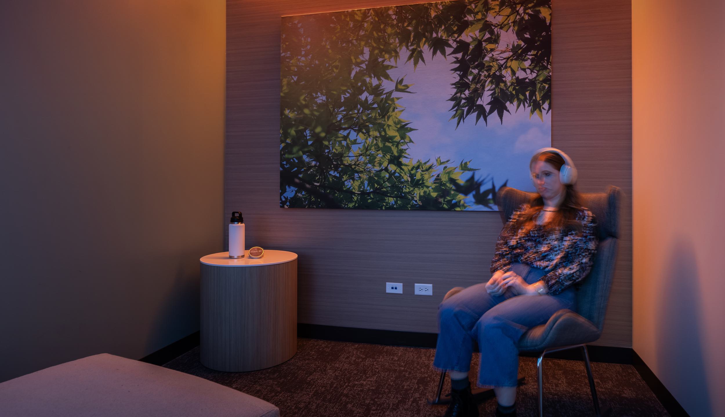 A person sits in a cozy room wearing headphones, surrounded by warm lighting and a nature-themed wall art, exuding calmness and relaxation.