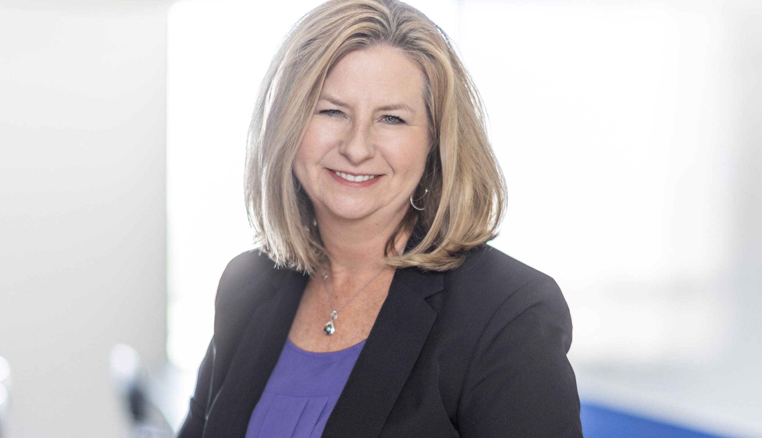 A smiling woman with shoulder-length blonde hair, wearing a black blazer over a purple blouse, poses in a bright, modern setting.