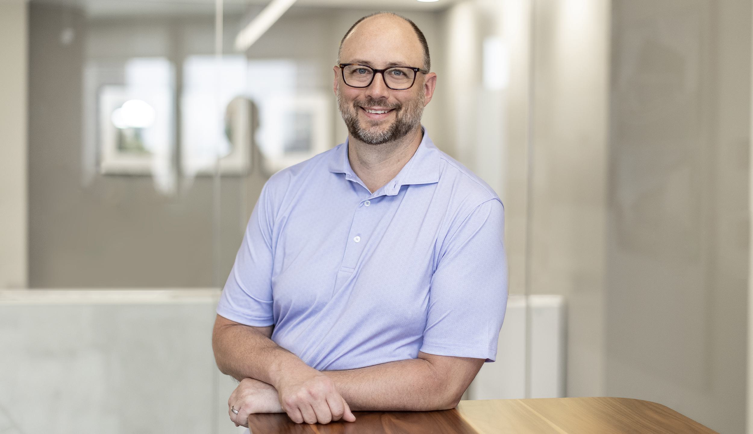 A smiling man in a light purple polo shirt leans on a wooden table, set against a modern, softly lit office background.
