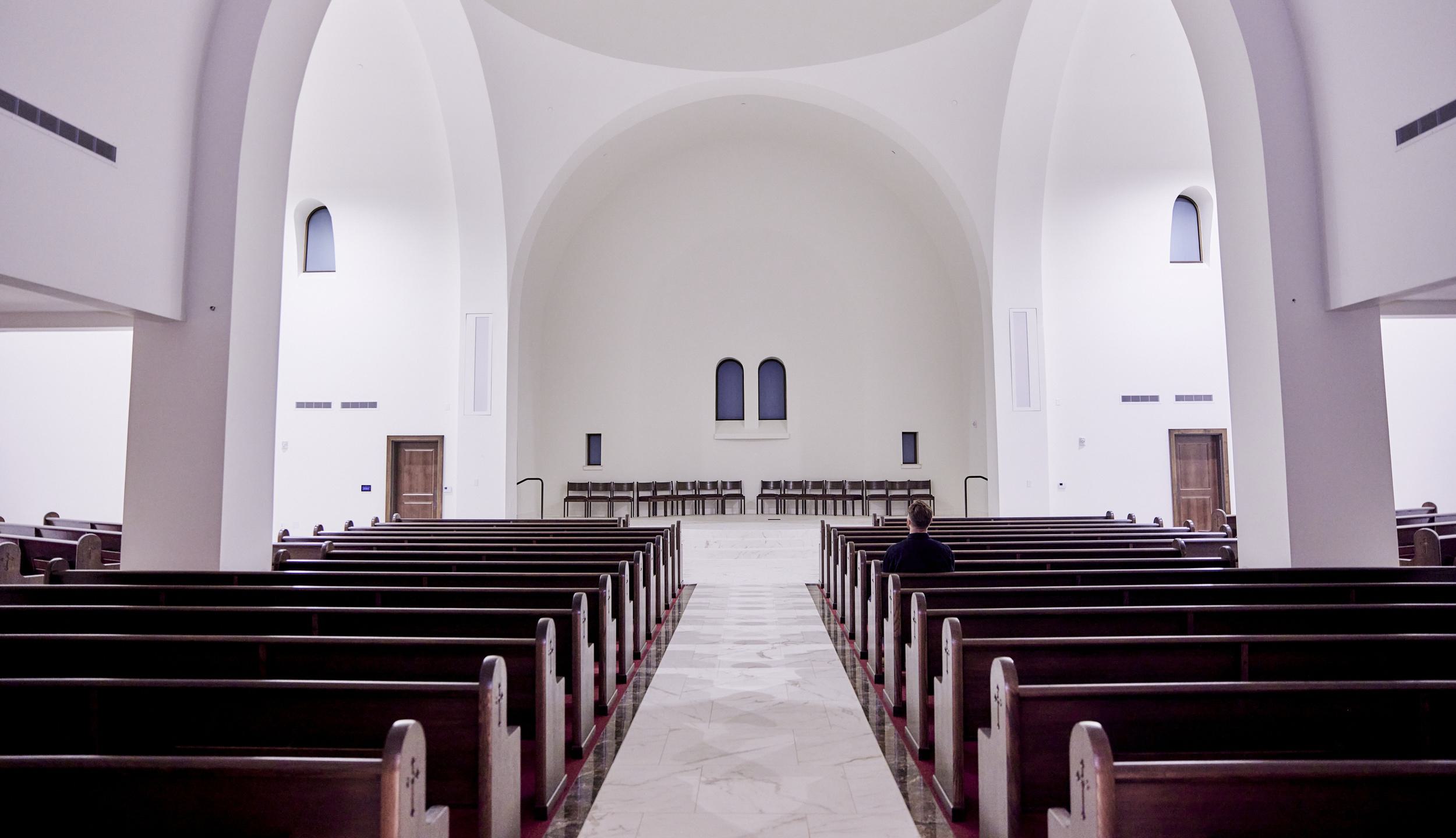 A spacious, modern interior of a church features long wooden pews, arched ceilings, and subtle lighting, creating a serene atmosphere.