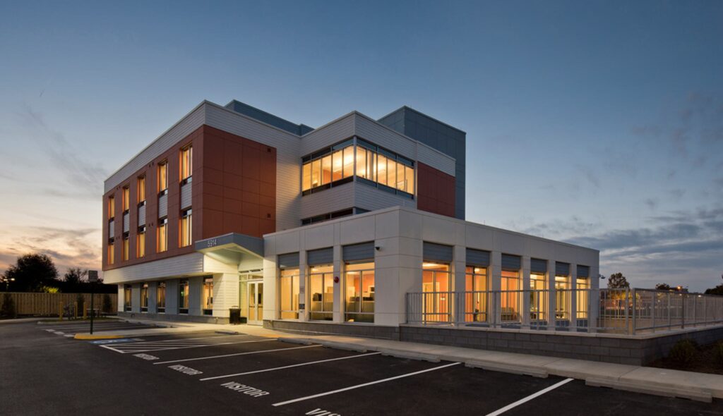 A modern multi-story building features large windows and a mix of red and gray exteriors, set against a twilight sky.