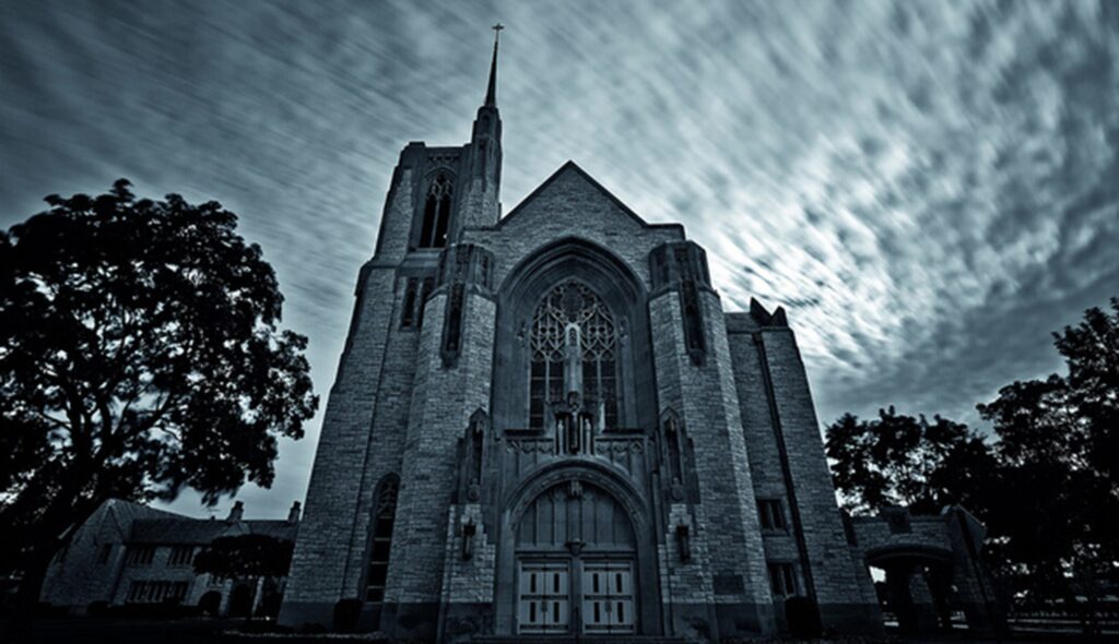 A grand, gothic-style church stands against a moody, cloud-filled sky, surrounded by dark trees, creating an eerie yet striking atmosphere.