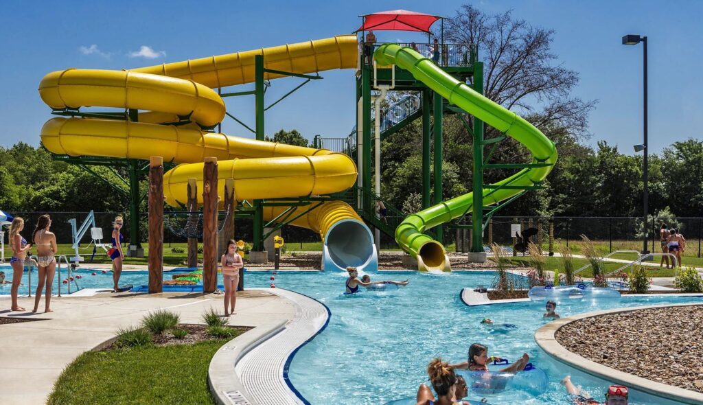 A vibrant water park features soaring yellow and green slides, with many children enjoying the nearby lazy river and splashing in the pool.