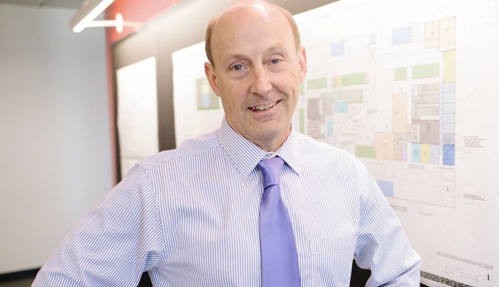 A man in a striped shirt and tie smiles confidently, standing in front of a detailed planning diagram in a modern office setting.