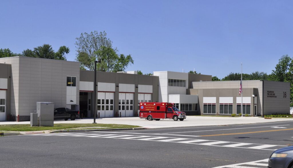 A modern fire station features large garage doors, an ambulance parked out front, and landscaped grounds under a clear blue sky.