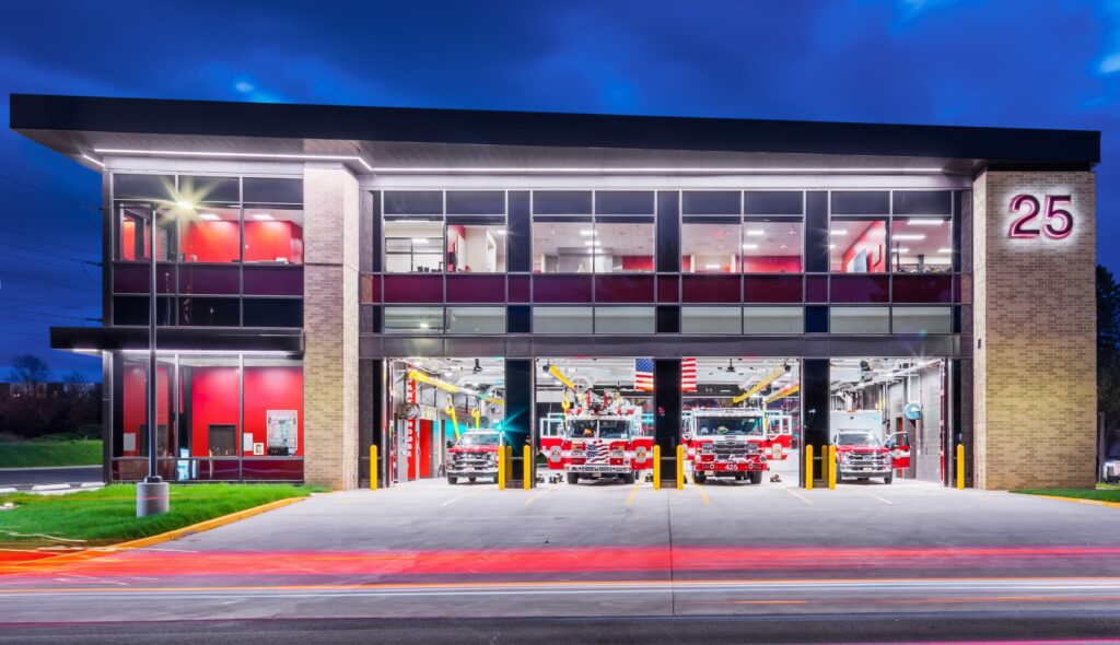 A modern fire station with large windows and multiple fire trucks visible. The exterior features a sleek design, illuminated in twilight.