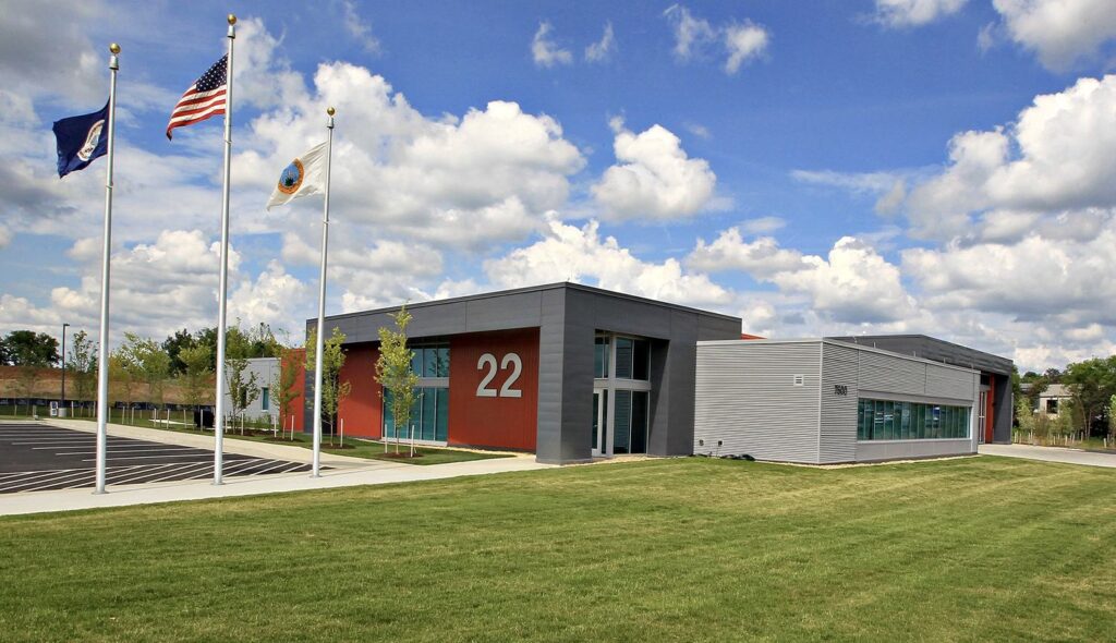 A modern building features large windows, a prominent number 22, and is flanked by flagpoles under a partly cloudy blue sky.