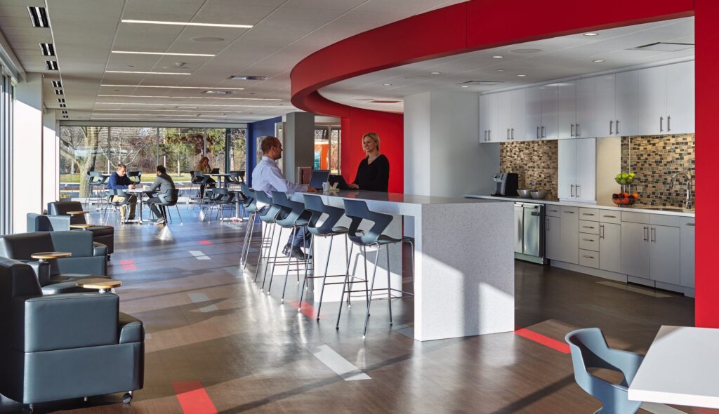A modern, brightly lit workspace features a café area with bar stools, a kitchen, and several seating arrangements for collaboration.