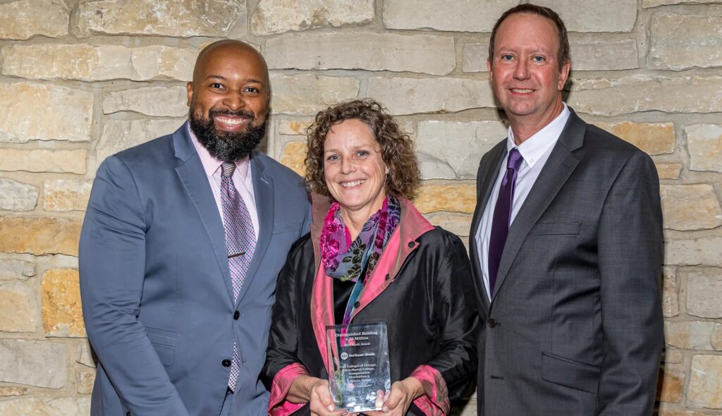 Three individuals smile together, with a woman holding an award, set against a stone wall background.