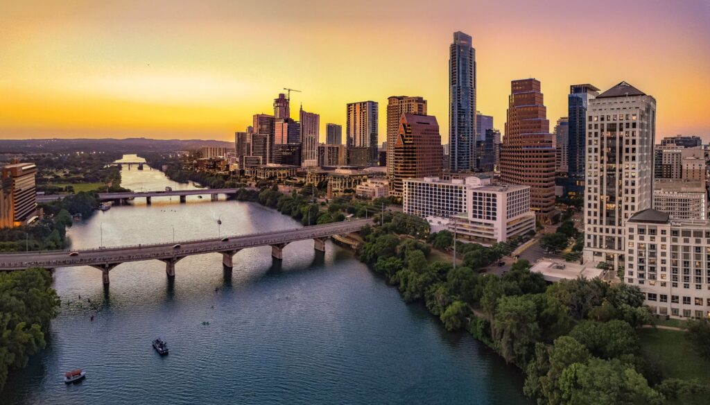 A vibrant city skyline at sunset, reflecting on a river, with lush greenery and a bridge connecting both sides.
