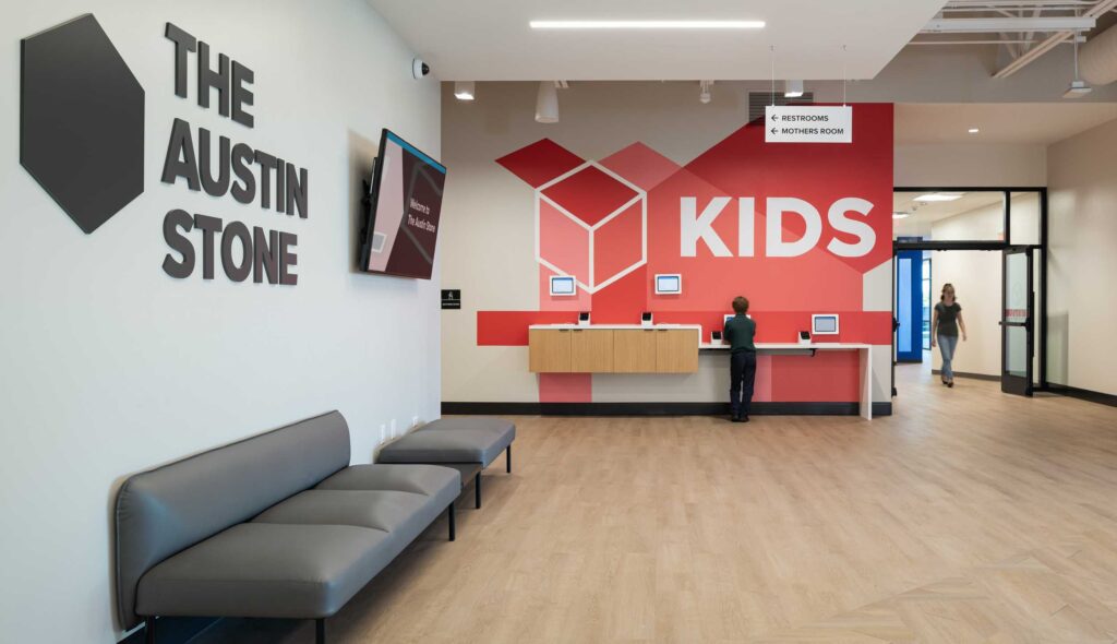 A bright lobby at The Austin Stone South church features a welcoming reception area with a bold red and white "KIDS" wayfinding graphic with sign-in kiosks and seating, designed for family registration.
