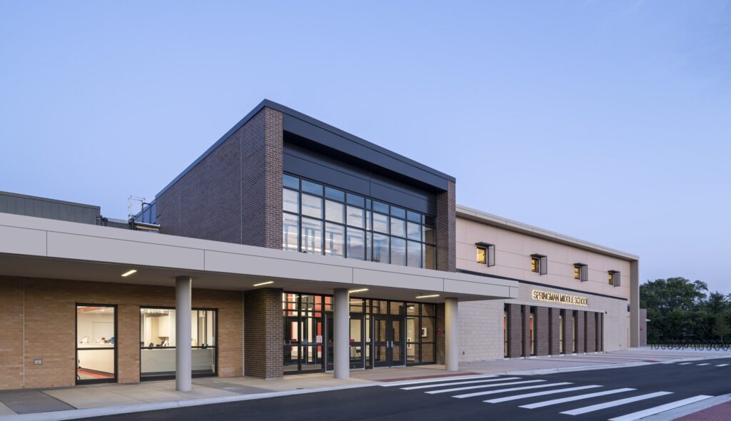 A modern school building features a large glass atrium, brick façade, and spacious entrance, set against a clear blue sky.