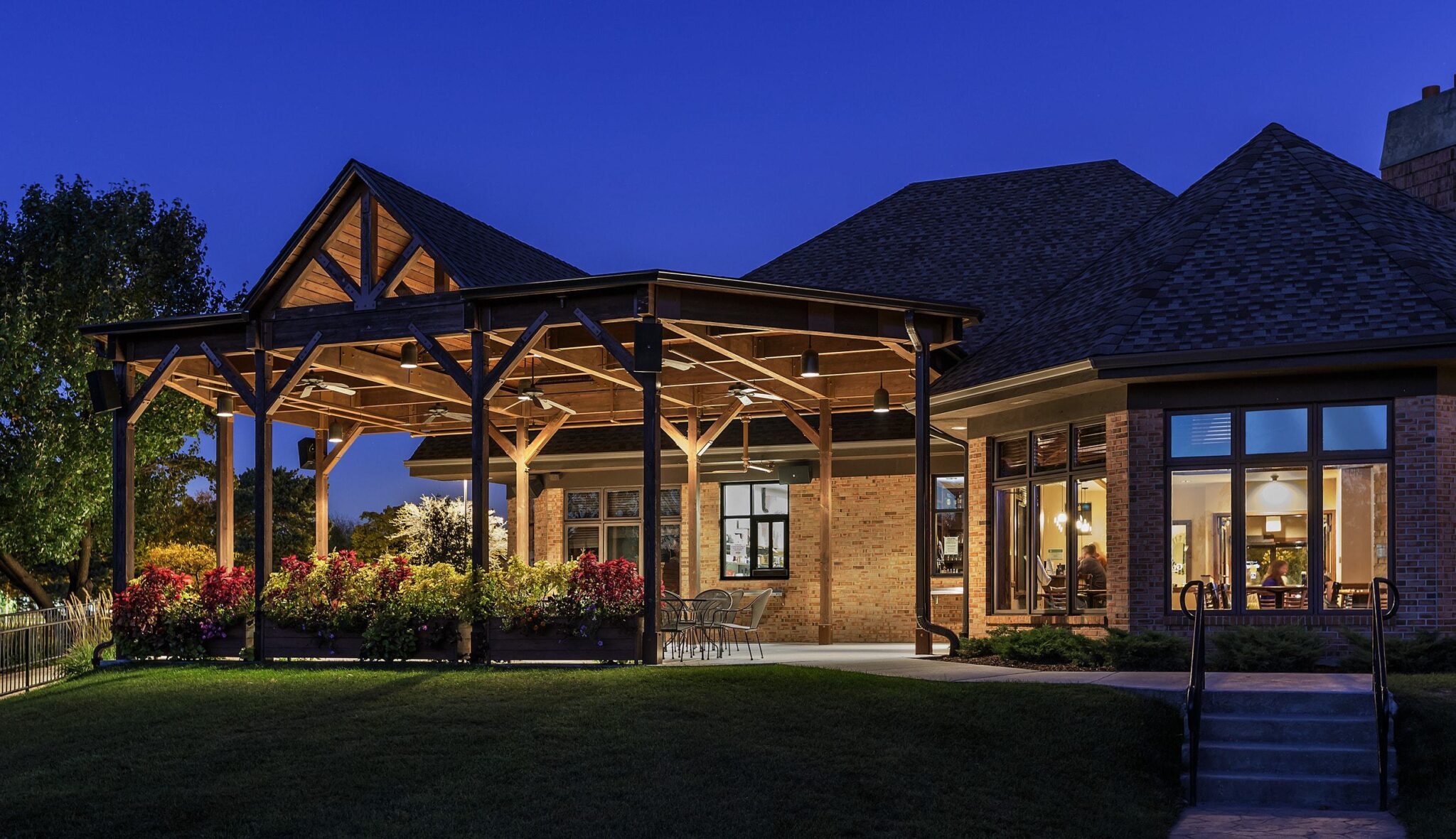 A modern house features a spacious covered patio with wooden beams, surrounded by colorful landscaping, illuminated in soft evening light.