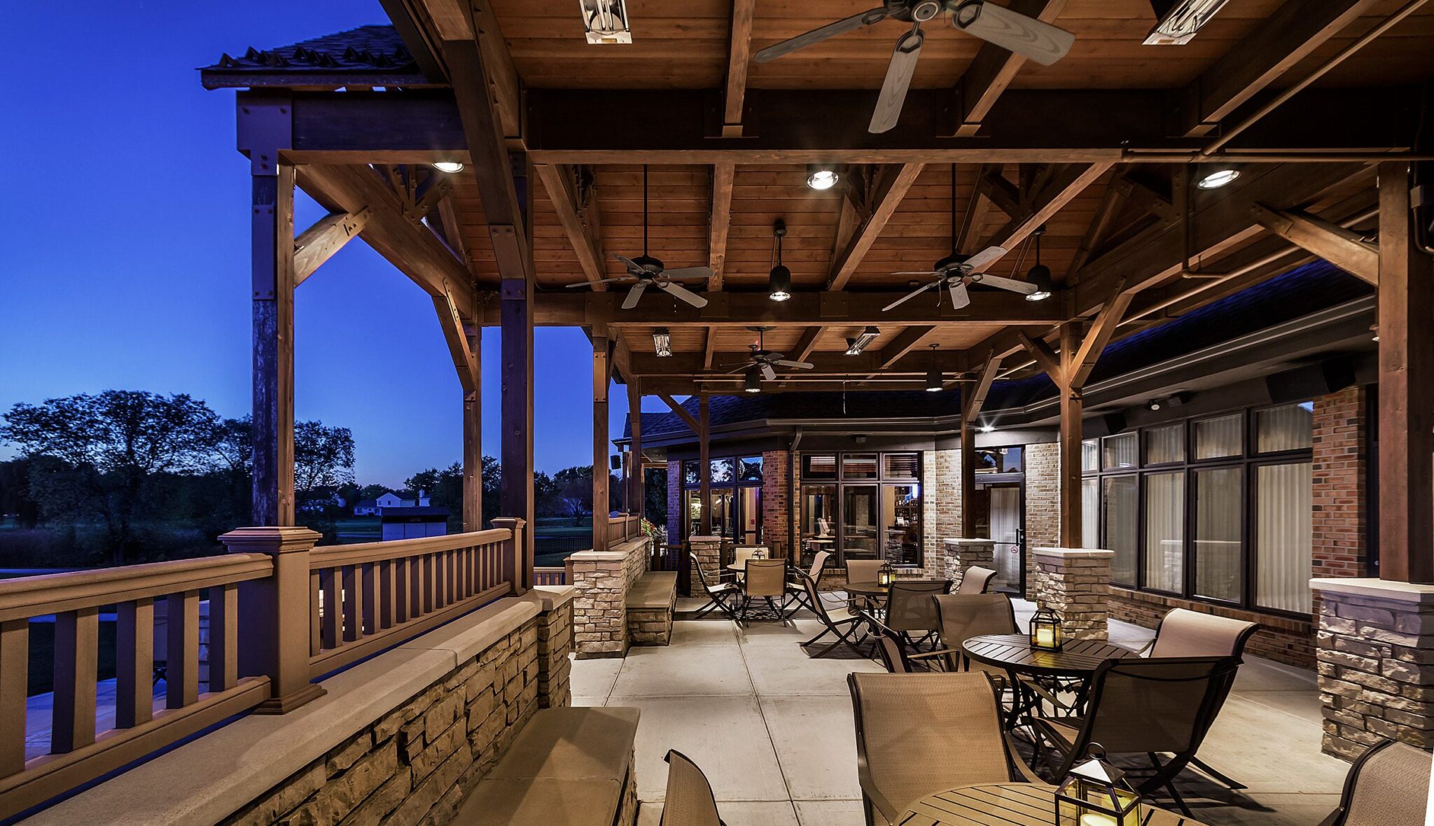 A cozy outdoor patio with wooden beams, stone accents, and comfortable seating, illuminated under a twilight sky. Fans gently circulate the air.