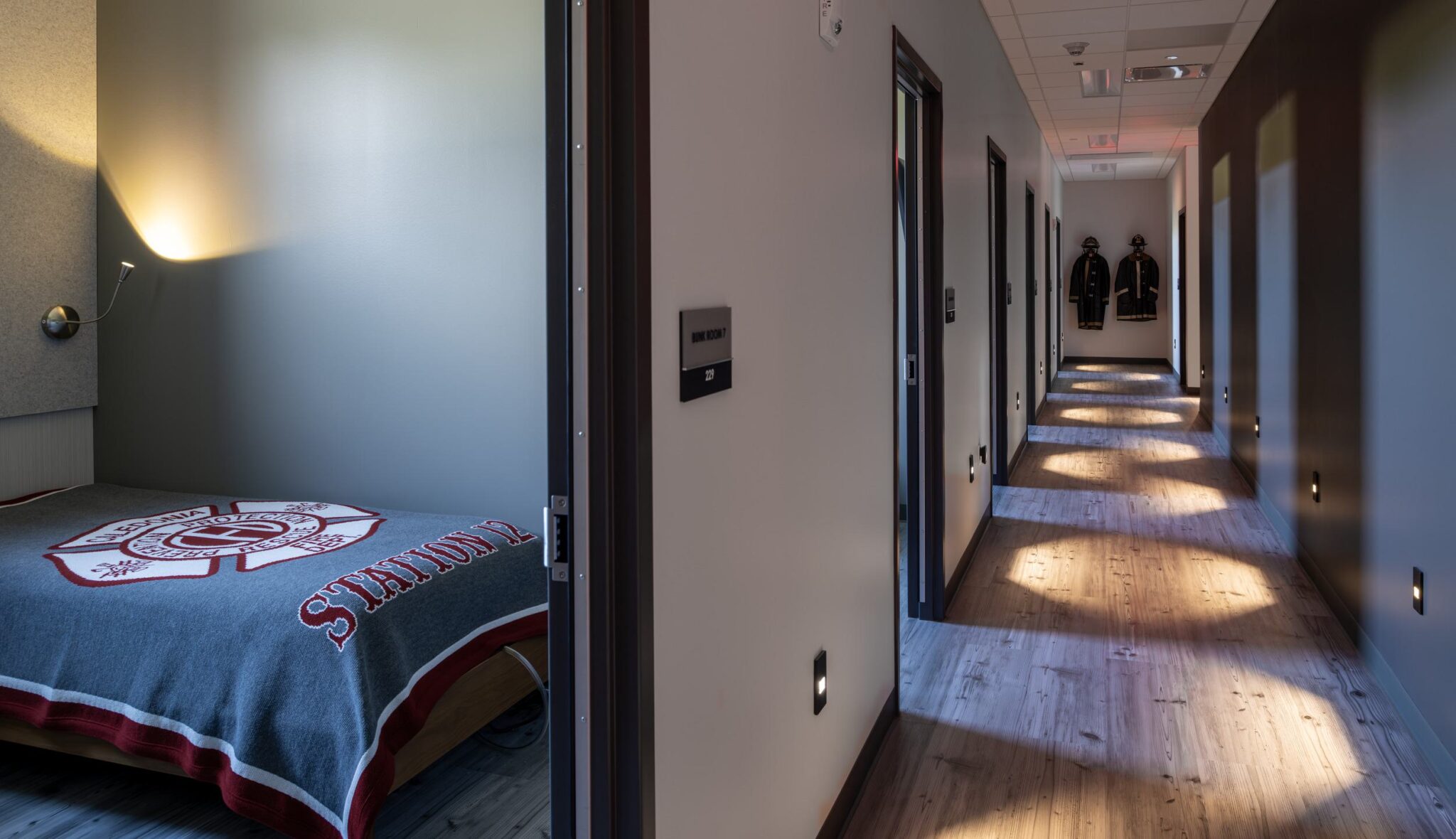 A well-lit hallway with wooden floors leads to a cozy room featuring a bed covered with a fire department-themed blanket and uniforms on the wall.