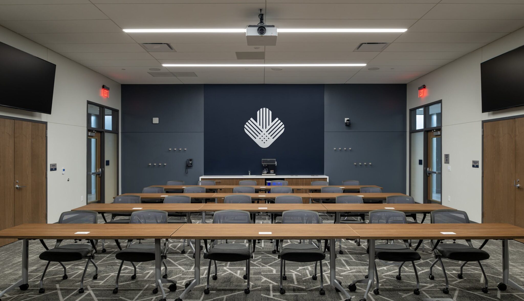 A modern conference room features rows of tables and chairs, with a projector and a logo displayed on the wall.